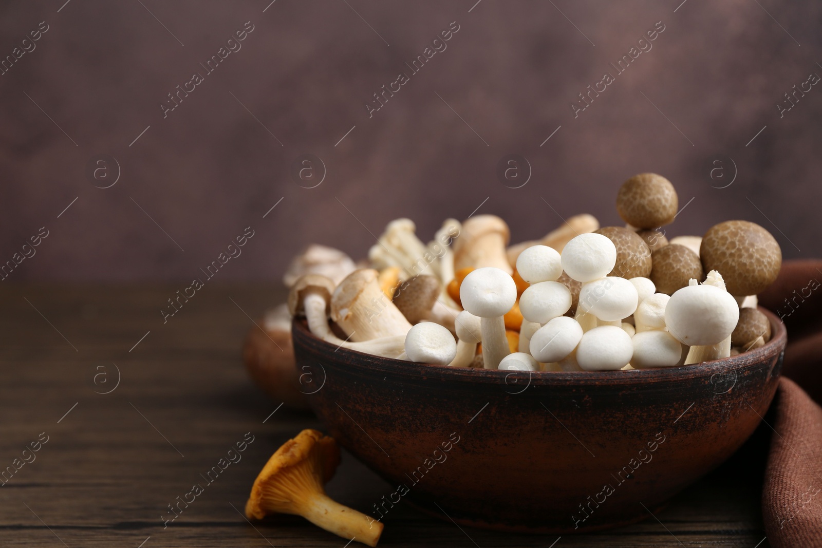 Different raw mushrooms in bowl on wooden table against brown background, closeup. Space for text Photo of Different raw mushrooms in bowl on wooden table against brown background, closeup. Space for text