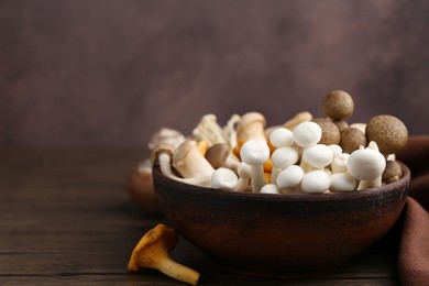 Photo of Different raw mushrooms in bowl on wooden table against brown background, closeup. Space for text