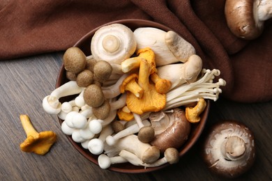 Photo of Different raw mushrooms in bowl on wooden table, flat lay