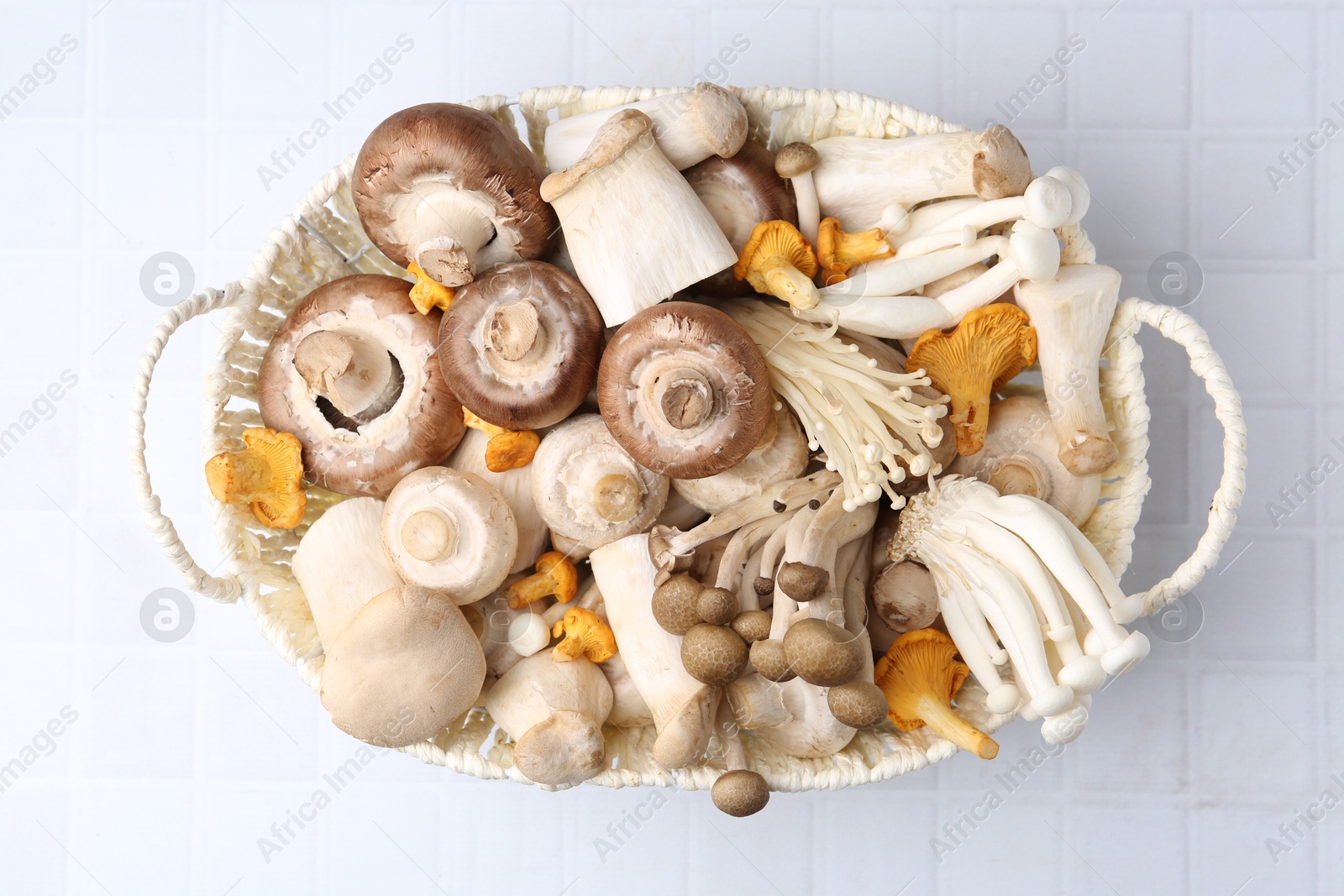 Photo of Different raw mushrooms in wicker basket on white tiled table, top view