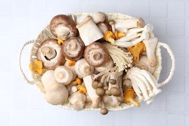 Photo of Different raw mushrooms in wicker basket on white tiled table, top view
