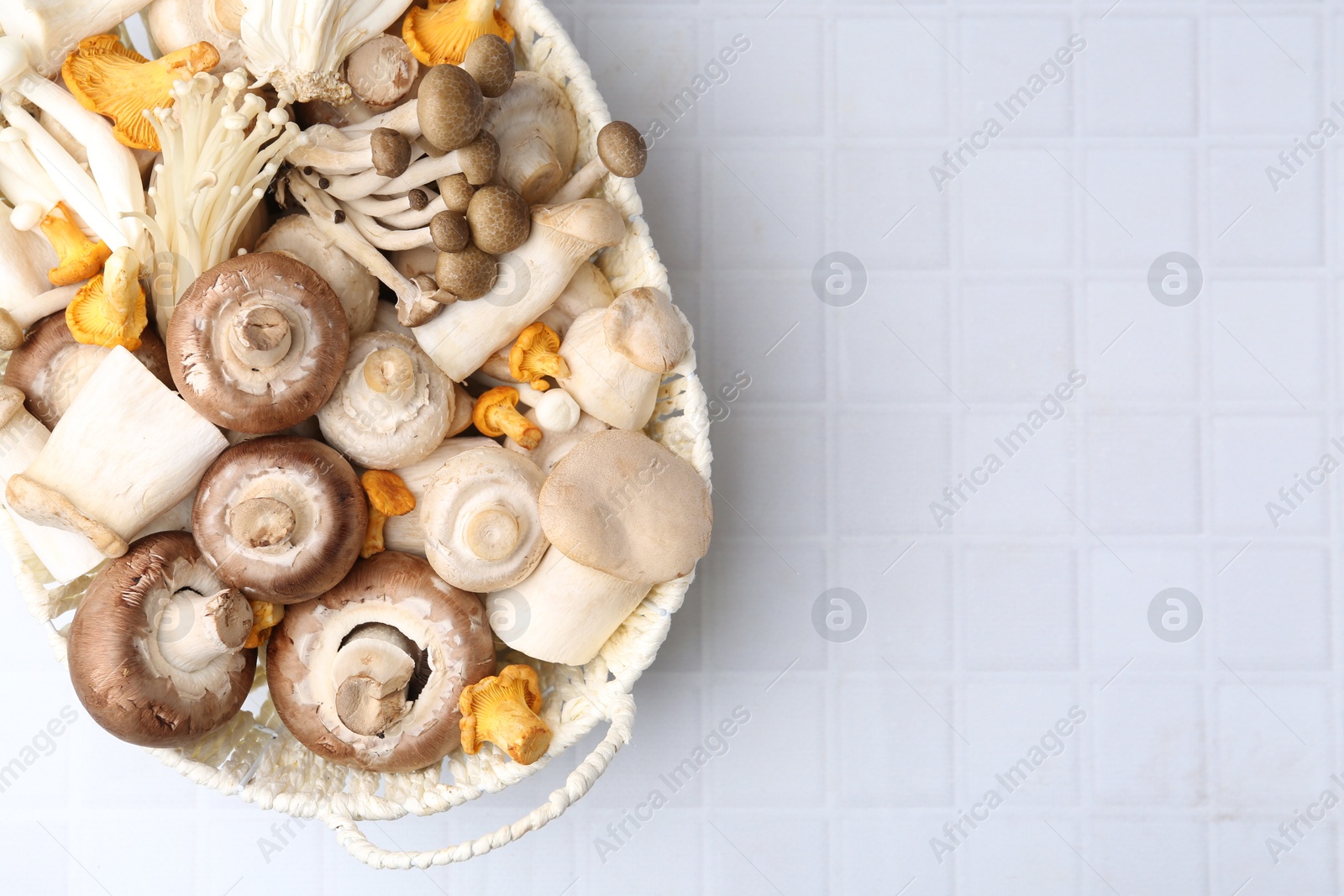 Photo of Different raw mushrooms in wicker basket on white tiled table, top view. Space for text