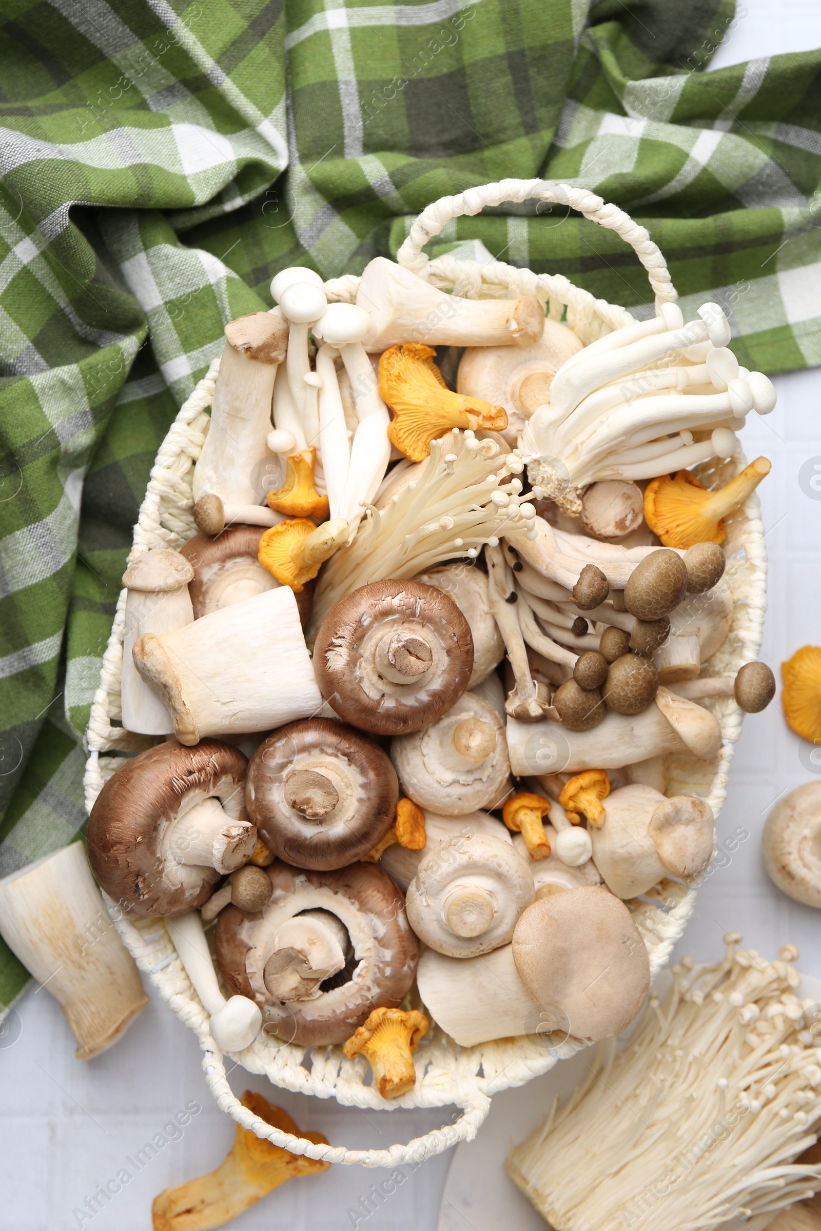 Photo of Different raw mushrooms in wicker basket on white tiled table, flat lay
