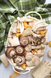 Photo of Different raw mushrooms in wicker basket on white tiled table, flat lay