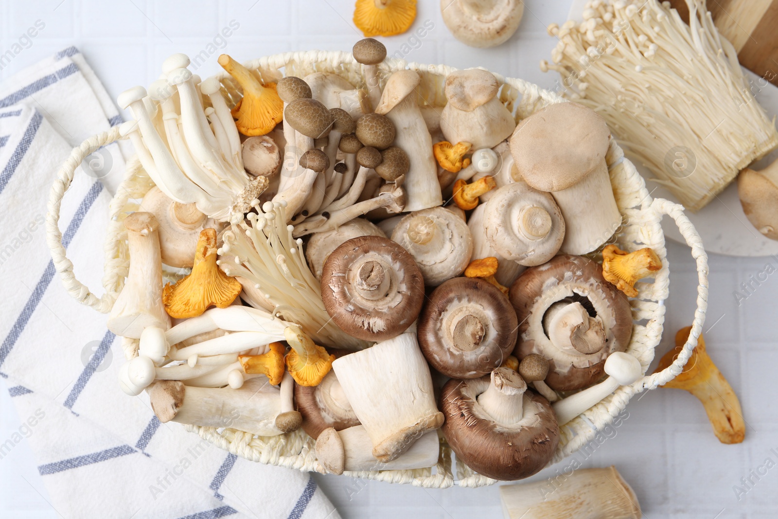 Photo of Different raw mushrooms in wicker basket on white tiled table, flat lay