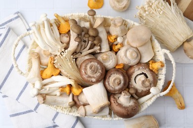 Photo of Different raw mushrooms in wicker basket on white tiled table, flat lay