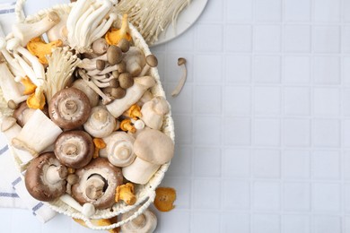 Photo of Different raw mushrooms in wicker basket on white tiled table, flat lay. Space for text