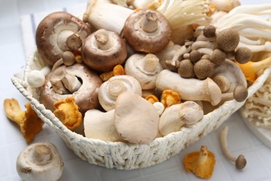 Photo of Different raw mushrooms in wicker basket on white tiled table, closeup