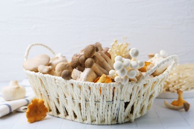 Photo of Different raw mushrooms in wicker basket on white tiled table, closeup