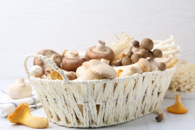 Photo of Different raw mushrooms in wicker basket on white tiled table, closeup