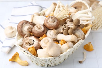 Photo of Different raw mushrooms in wicker basket on white tiled table, closeup
