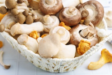 Photo of Different raw mushrooms in wicker basket on white tiled table, closeup