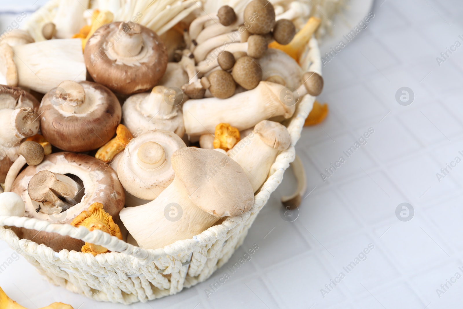 Photo of Different raw mushrooms in wicker basket on white tiled table, closeup