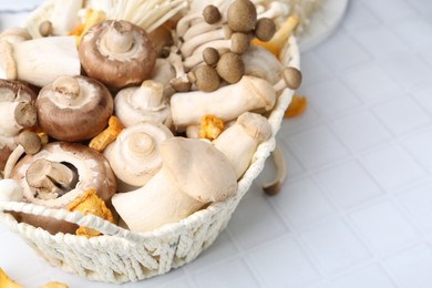 Photo of Different raw mushrooms in wicker basket on white tiled table, closeup