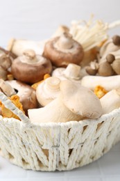 Photo of Different raw mushrooms in wicker basket on white tiled table, closeup