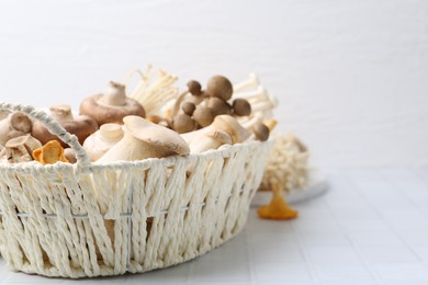 Photo of Different raw mushrooms in wicker basket on white tiled table, closeup. Space for text