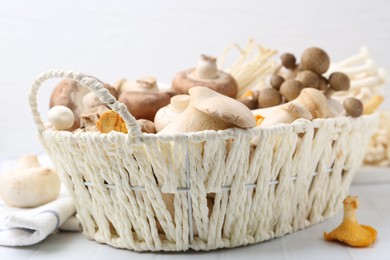 Photo of Different raw mushrooms in wicker basket on white tiled table, closeup