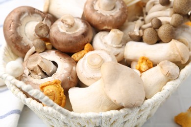 Photo of Different raw mushrooms in wicker basket on white table, closeup