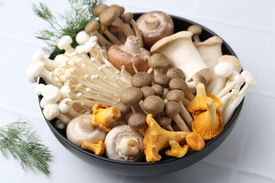 Photo of Different raw mushrooms in bowl and dill on white tiled table, closeup