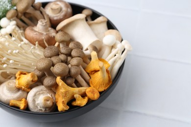 Photo of Different raw mushrooms in bowl on white tiled table, closeup. Space for text