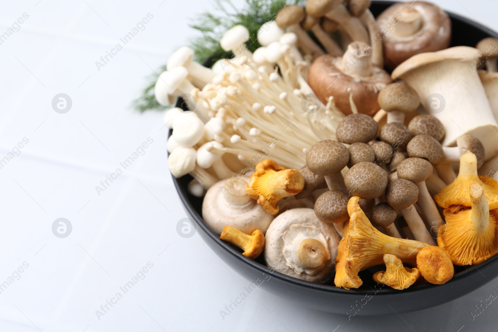 Photo of Different raw mushrooms in bowl on white tiled table, closeup. Space for text