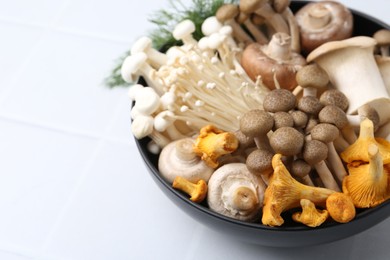 Photo of Different raw mushrooms in bowl on white tiled table, closeup. Space for text