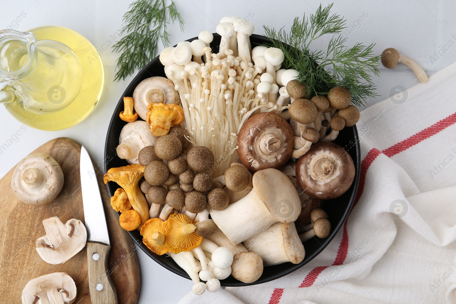 Photo of Different raw mushrooms in bowl, oil, knife and dill on white tiled table, flat lay