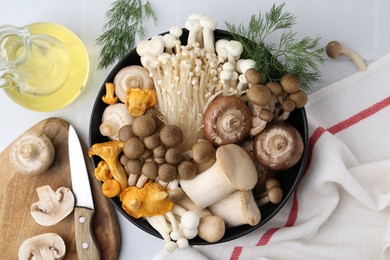 Photo of Different raw mushrooms in bowl, oil, knife and dill on white tiled table, flat lay