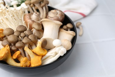 Photo of Different raw mushrooms in bowl on white tiled table, closeup. Space for text
