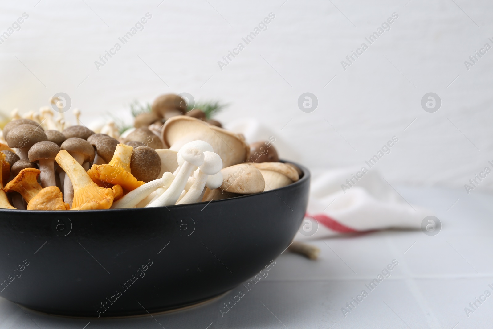 Photo of Different raw mushrooms in bowl on white tiled table, closeup. Space for text