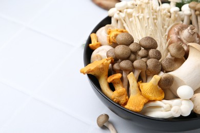 Photo of Different raw mushrooms in bowl on white tiled table, closeup. Space for text
