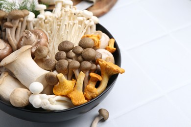 Photo of Different raw mushrooms in bowl on white tiled table, closeup. Space for text