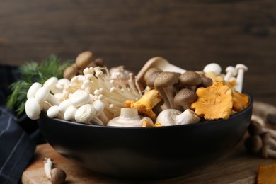 Photo of Different raw mushrooms in bowl on table against wooden background, closeup
