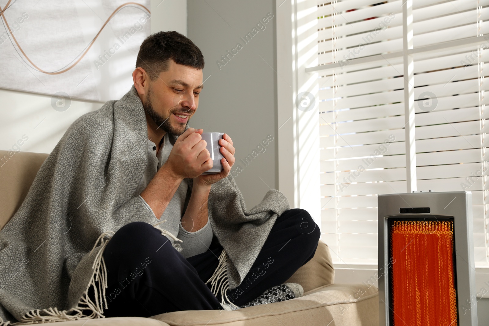 Man with cup of hot drink near electric heater at home Image of Man with cup of hot drink near electric heater at home