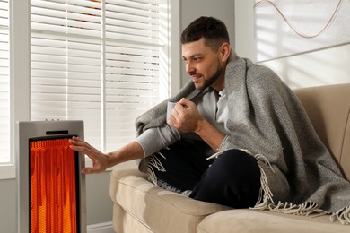 Image of Man with cup of hot drink near electric heater at home