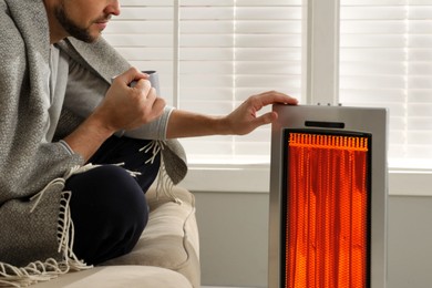Image of Man with cup of hot drink near electric heater at home, closeup