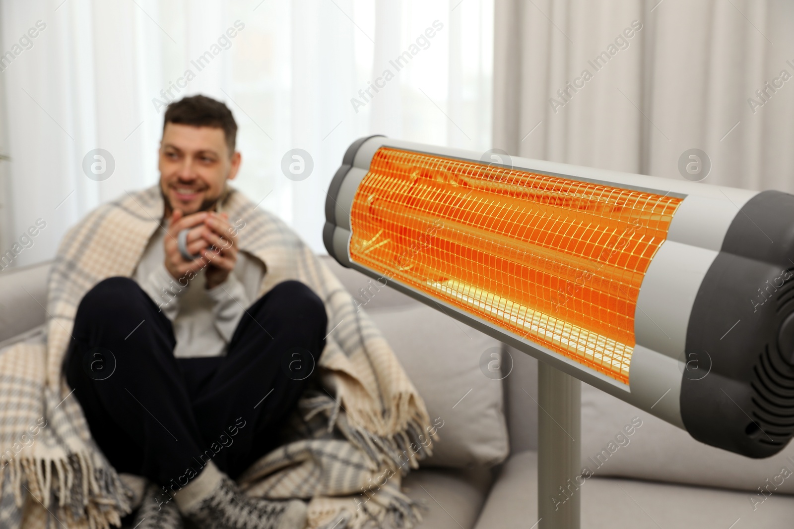 Man with cup of hot drink near electric heater at home, selective focus Image of Man with cup of hot drink near electric heater at home, selective focus