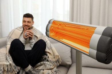 Image of Man with cup of hot drink near electric heater at home, selective focus