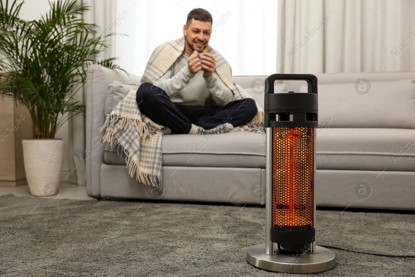 Man with cup of hot drink near electric heater at home, selective focus Image of Man with cup of hot drink near electric heater at home, selective focus
