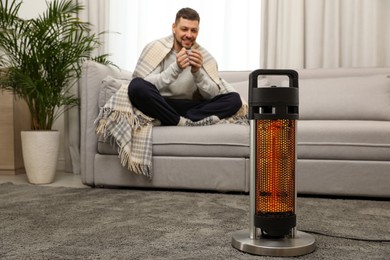 Image of Man with cup of hot drink near electric heater at home, selective focus