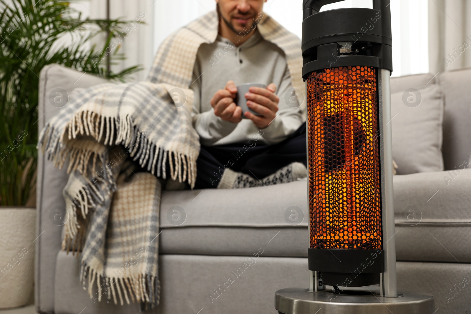 Man with cup of hot drink near electric heater at home, selective focus Image of Man with cup of hot drink near electric heater at home, selective focus