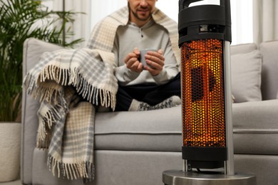 Image of Man with cup of hot drink near electric heater at home, selective focus