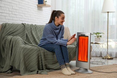 Image of Woman warming her hands near electric heater at home