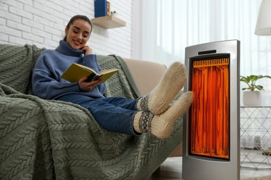 Image of Woman warming her feet near electric heater while reading book on sofa at home