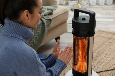 Image of Woman warming her hands near electric heater at home