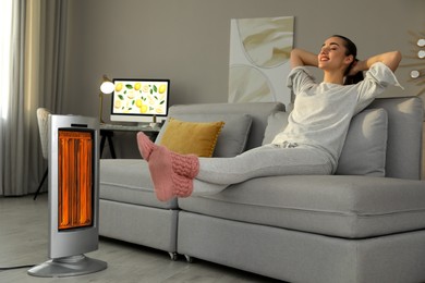 Image of Woman warming her feet near electric heater on sofa at home