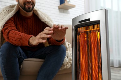 Image of Man warming his hands near electric heater at home, closeup