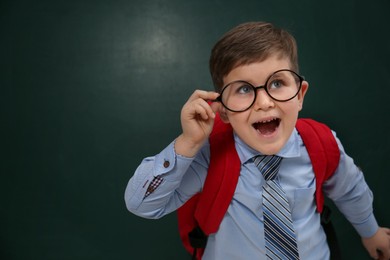 Photo of Funny little child wearing glasses near chalkboard, space for text. First time at school