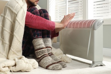 Photo of Man warming hands near electric heater at home