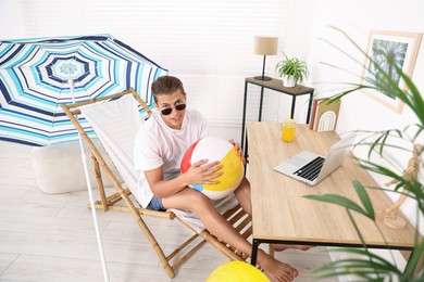 Photo of Happy man in sunglasses with beach ball having fun on sunbed at table indoors. Dreaming about vacation at sea